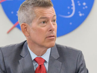 a man with gray hair and pale complexion wears a gray suit and red tie while sitting at a table under a red, white and blue NASA logo on the wall behind him