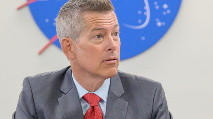 a man with gray hair and pale complexion wears a gray suit and red tie while sitting at a table under a red, white and blue NASA logo on the wall behind him