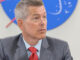 a man with gray hair and pale complexion wears a gray suit and red tie while sitting at a table under a red, white and blue NASA logo on the wall behind him