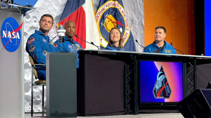 Three men and a woman, all in blue flight suits, sit at a desk during a press conference