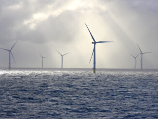 A series of tall wind turbines in the ocean, illuminated by beams of sunlight filtering through baps of heavy clouds.
