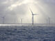 A series of tall wind turbines in the ocean, illuminated by beams of sunlight filtering through baps of heavy clouds.