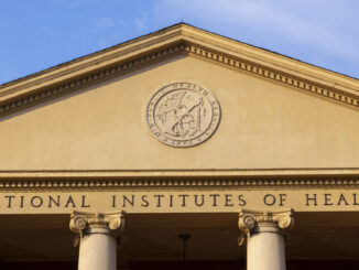 A view of the exterior of a building with a triangular top supported by pillars. Beneath a circular logo are the words "national Institutes of Health"