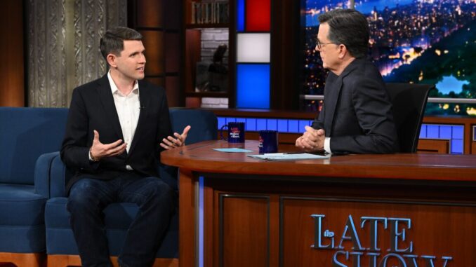 Stephen Colbert interviews a guest while sitting at his desk on the set of his late-night show.