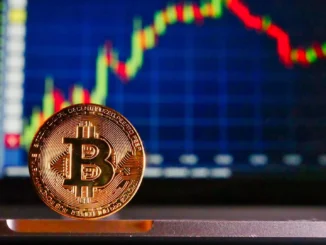 A physical Bitcoin coin in the foreground with a financial candlestick chart on a computer screen in the background.
